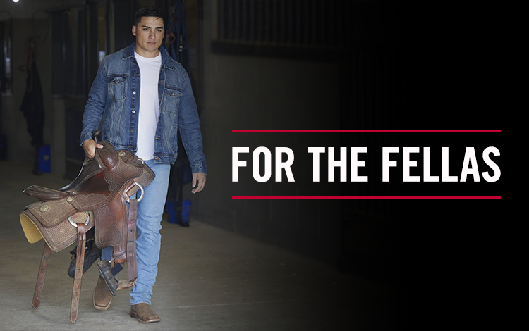 For The Fellas. A man wearing a denim jacket and carrying a saddle walking down a barn. Shop Men.